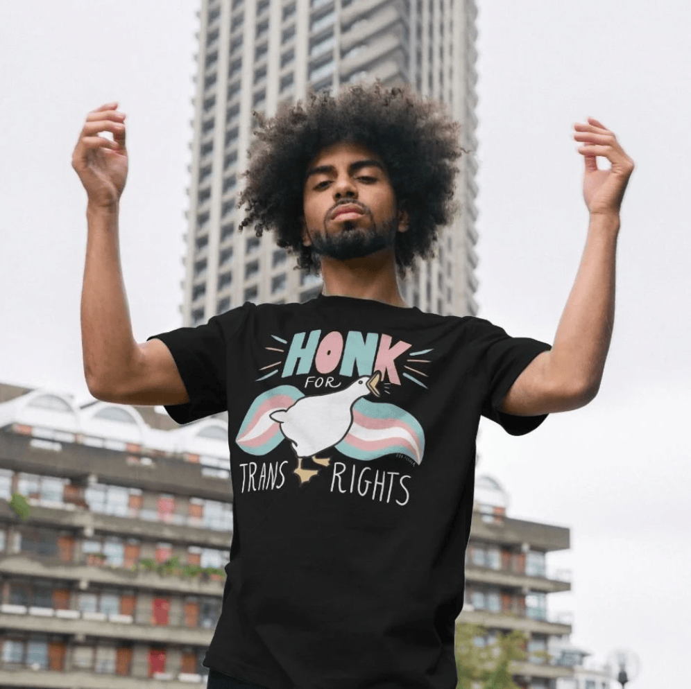 Someone with an afro standing in front of a tower block with both arms raised, wearing the black Honk For Trans Rights t-shirt.