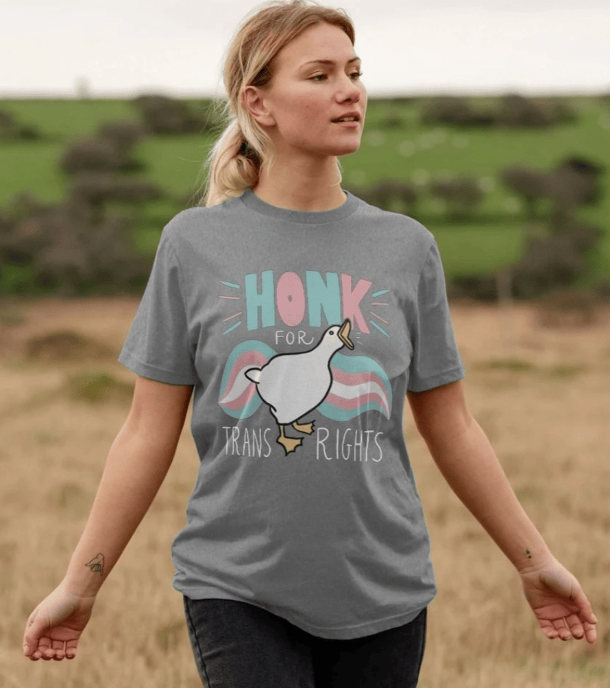 Someone with a blonde ponytail walking through a grassy field, wearing the grey marl Honk For Trans Rights t-shirt.