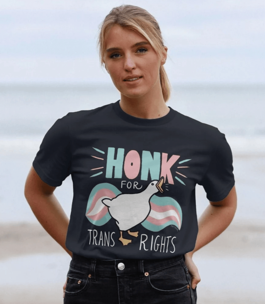 Someone with blonde hair standing on a beach with the sea behind them, wearing the navy Honk For Trans Rights t-shirt tucked into black jeans.